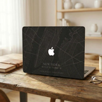 Personalized City Map MacBook case from TrioAuraDesign, shown on a rustic wooden desk in a warm home office. The dark hard shell features a custom New York street map, city name, coordinates, the phrase “YOUR PLACE. YOUR STORY,” and the Apple logo visible at center. Lifestyle view of a custom map laptop cover for compatible MacBook Air, Pro, and Neo models.