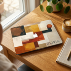 Personalized abstract geometric MacBook case with custom name Olivia in terracotta mustard navy and cream color blocks, displayed on wooden desk near sunny window with coffee cup, potted plant and linen notebook — bold architectural collage hard shell laptop cover with elegant serif name personalization, Apple logo visible through an unprinted area, slim snap-on fit, for MacBook Air 13 M2 M3 M4 M5 and MacBook Pro 14 16 M3 M4 M5, modern aesthetic personalized gift for creative professionals