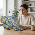Lifestyle photo of a woman sitting at a light wooden table, using a MacBook. The laptop is protected by a hardshell case featuring a stunning, colorful stained glass mosaic design. The artwork portrays hummingbirds and lilies with golden outlines against a blended blue, purple, and yellow mosaic background. The woman smiles as she works, with a blurred office background, potted plants, and a window visible.