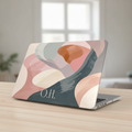 A MacBook with a personalized neutral abstract case in soft pink, sage green, and beige on a wooden desk with a window view, featuring the monogram O.H.
