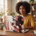 A personalized MacBook case on a wooden desk with a student using it, featuring a vibrant portrait of a Black woman with coral and pink flowers, and the custom typography SOPHIA MUSE on a neutral beige background.