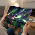 Close-up lifestyle photo of a person's hands holding an open MacBook. The lid is protected by a hardshell case featuring a vivid Northern Lights design, with emerald green and purple Aurora Borealis glowing over an arctic landscape and a starry night sky.