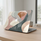 A MacBook with a personalized neutral abstract case in soft pink, sage green, and beige on a wooden desk with a window view, featuring the monogram O.H.
