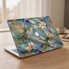 Lifestyle photo of a MacBook on a light wooden table by a window. The laptop is protected by a hardshell case featuring a detailed stained glass mosaic design of emerald green hummingbirds and lilies with gold-accented outlines. A partial cutout in the center of the design reveals the Apple logo on the silver laptop lid.