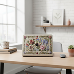 A MacBook with a decorative vintage floral hard shell case sitting on a light wooden desk in a bright, modern office. The case features a detailed 3D-effect botanical design with roses, wisteria, and lilies on a linen-textured background. The workspace is neatly arranged with a coffee mug, books, a wireless mouse, and a small succulent plant.