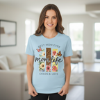 A happy woman posing in a modern living room, wearing a light blue cotton "Mom Life: Chaos & Love" t-shirt with the stylish leopard floral butterfly print.