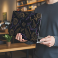 Lifestyle photo of a person carrying a MacBook with two hands. The glossy hard shell case features a paleo-futuristic 3D design of golden fossil ammonites, ferns, and bones accented by glowing neon purple and cyan cyber-lines.