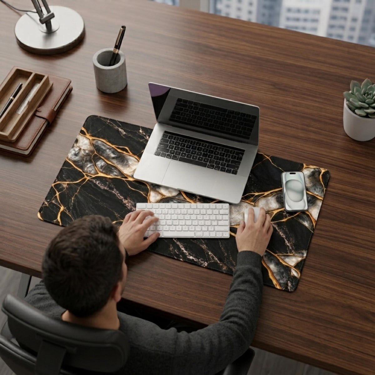 Top view of a man typing at a desk on the black marble and gold desk mat, with a laptop, white keyboard and phone resting on the mat — a black and gold workspace setup.