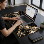 Woman working on a laptop resting on the black marble desk mat with gold kintsugi effect veins, on a dark desk with a notebook and plant.