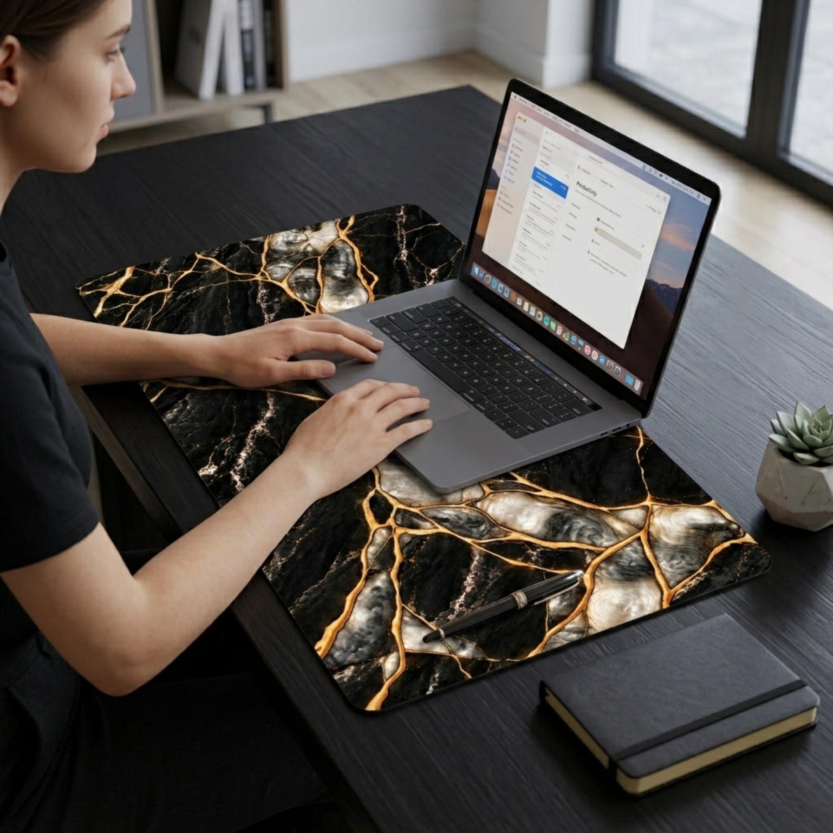 Woman working on a laptop resting on the black marble desk mat with gold kintsugi effect veins, on a dark desk with a notebook and plant.