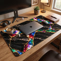 Jewel Tone style black marble desk mat on a wooden desk with a closed MacBook, coffee cup, headphones and notebook. Black stone with gold kintsugi effect veins and glowing emerald, sapphire, ruby and violet opal-like stones, printed on a soft fabric surface.