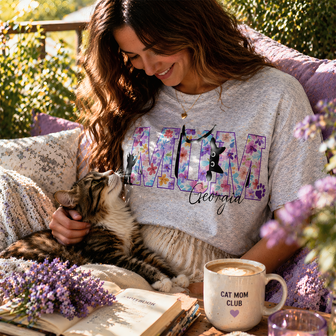 Lifestyle photo of a smiling woman cuddling her tabby cat, wearing a personalized grey 'Cat Mum' T-shirt with purple floral lettering and the name 'Georgia', in a cozy sunlit garden setting with lavender.