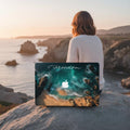 Woman sitting on coastal rocks at sunset holding MacBook with custom ocean aerial photography case, Freedom personalized laptop cover