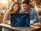 Custom initial MacBook case with dark navy botanical floral design, personalized with white script initials J.L., lifestyle photo of smiling couple at outdoor cafe table with coffee and croissant