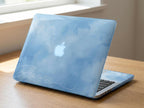 Custom name MacBook case with soft blue watercolor sky and cloud print, personalized with white signature script name Aurora, side angle view on wooden desk by window