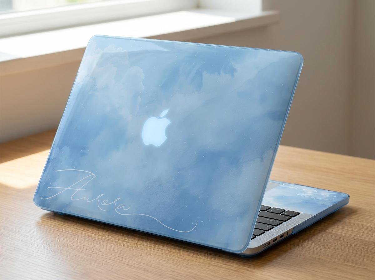 Custom name MacBook case with soft blue watercolor sky and cloud print, personalized with white signature script name Aurora, side angle view on wooden desk by window