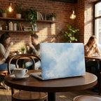 Custom name MacBook case with soft blue watercolor sky and cloud print, lifestyle photo on cafe table with coffee cup and brick wall background