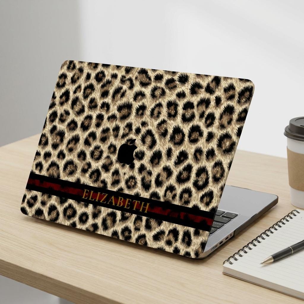 Custom leopard print MacBook case with personalized name in gold serif on dark stripe, cheetah pattern laptop cover, desk mockup with notebook and coffee