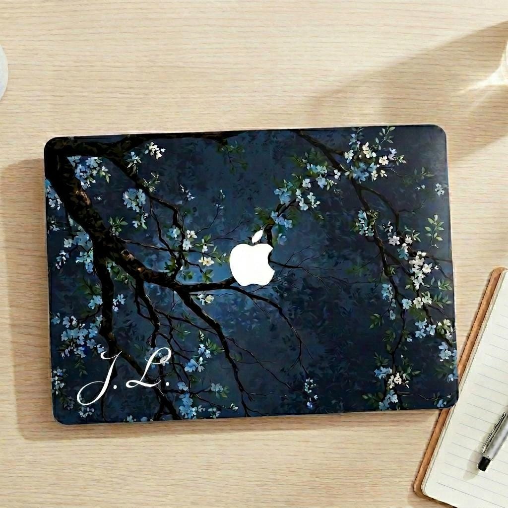 Custom initial MacBook case with dark navy botanical floral design, personalized with white script initials J.L., flat lay top view on wooden desk with notebook and pen