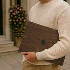 Man in a cream knit sweater holding a closed MacBook with a personalized wood-look case showing a warm walnut wood-grain print and custom text, with a blooming rose wall in the background. The wood grain and lettering are a high-definition UV print on a smooth polycarbonate hard shell, not real wood. A handsome personalized Father's Day or birthday gift for him, carried on the go, customizable with your own word and year for the dad, grandpa, or man who has everything.