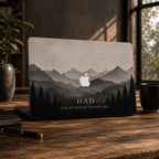 Personalized misty mountains MacBook case showing layered mountain peaks fading into fog above a silhouette pine forest in soft grey and charcoal tones, with the custom text DAD Our Adventure Started 2020 in elegant lettering. The Apple logo glows softly among the peaks. Shown on a partially open laptop on a dark wooden desk with a plant and notebook. The design and lettering are a high-definition UV print on a smooth polycarbonate hard shell. A personalized Father's Day adventure gift for him.
