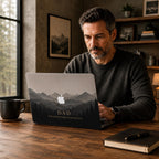 Man working on a laptop with a personalized misty mountains MacBook case showing the custom text DAD Our Adventure Started 2020, in a cozy home office with a bookshelf and mountain wall art. Layered mountain peaks fading into fog above a silhouette pine forest in soft grey and charcoal tones, the Apple logo glowing among the peaks. The design is a high-definition UV print on a smooth polycarbonate hard shell. A personalized Father's Day adventure gift for dad, customizable with your own words.