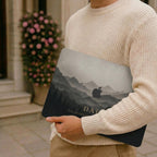 Man in a cream knit sweater holding a closed MacBook with a personalized misty mountains case, showing layered mountain peaks fading into fog above a pine forest in soft grey and charcoal tones with the custom text DAD and the Apple logo among the peaks, with a blooming rose wall in the background. The design is a high-definition UV print on a smooth polycarbonate hard shell. A personalized Father's Day adventure gift for him, carried on the go, customizable with your own words.
