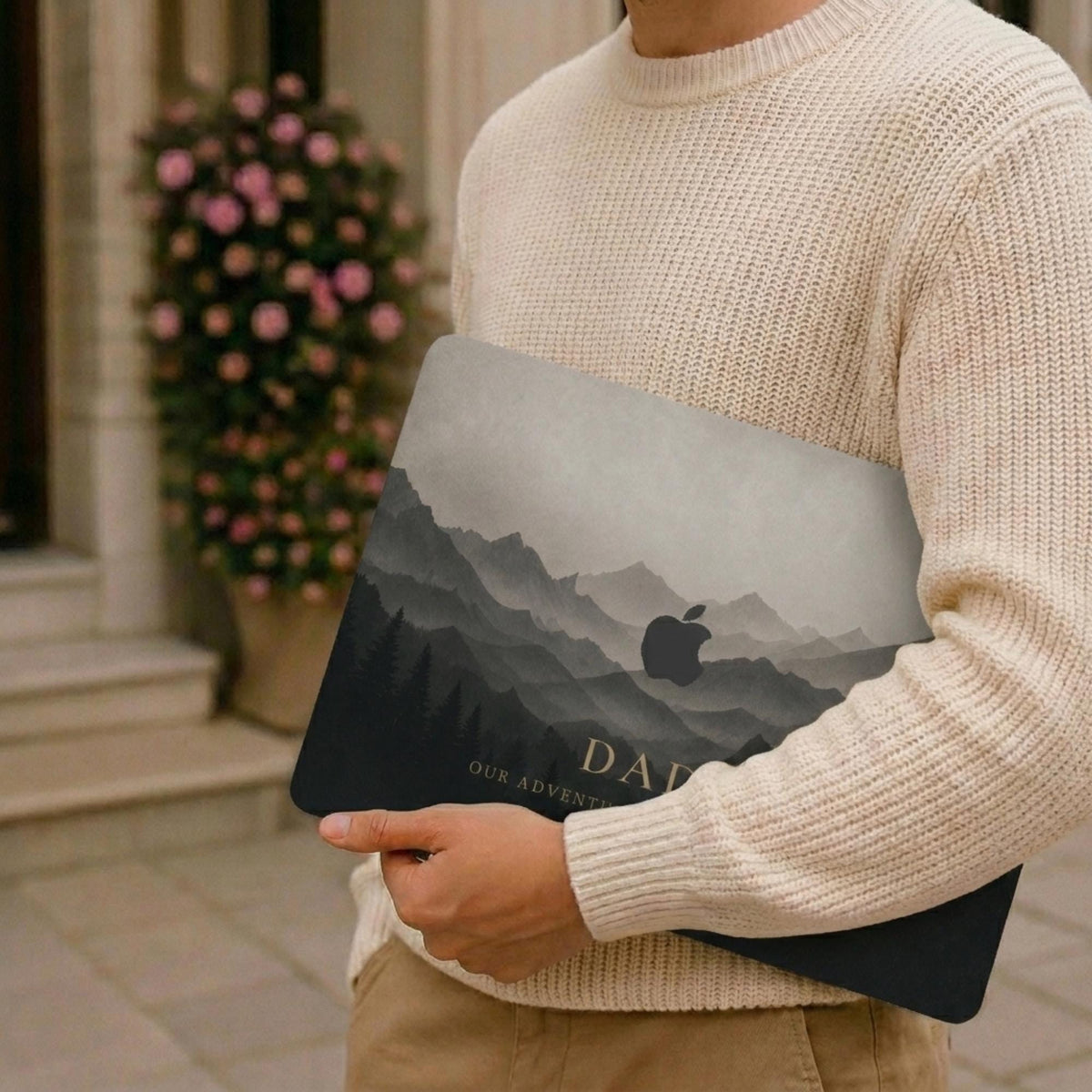 Man in a cream knit sweater holding a closed MacBook with a personalized misty mountains case, showing layered mountain peaks fading into fog above a pine forest in soft grey and charcoal tones with the custom text DAD and the Apple logo among the peaks, with a blooming rose wall in the background. The design is a high-definition UV print on a smooth polycarbonate hard shell. A personalized Father's Day adventure gift for him, carried on the go, customizable with your own words.