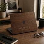 Personalized wood-look MacBook case with a warm walnut wood-grain print and the custom text GRANDPA Est. 1998 in elegant serif lettering, on a partially open laptop on a wooden desk with plants, books, glasses, and a coffee mug. The wood grain and lettering are a high-definition UV print on a smooth polycarbonate hard shell, not real wood. A heartfelt personalized gift for grandpa, Father's Day, or a milestone, fully customizable with your own word and year for any father figure.