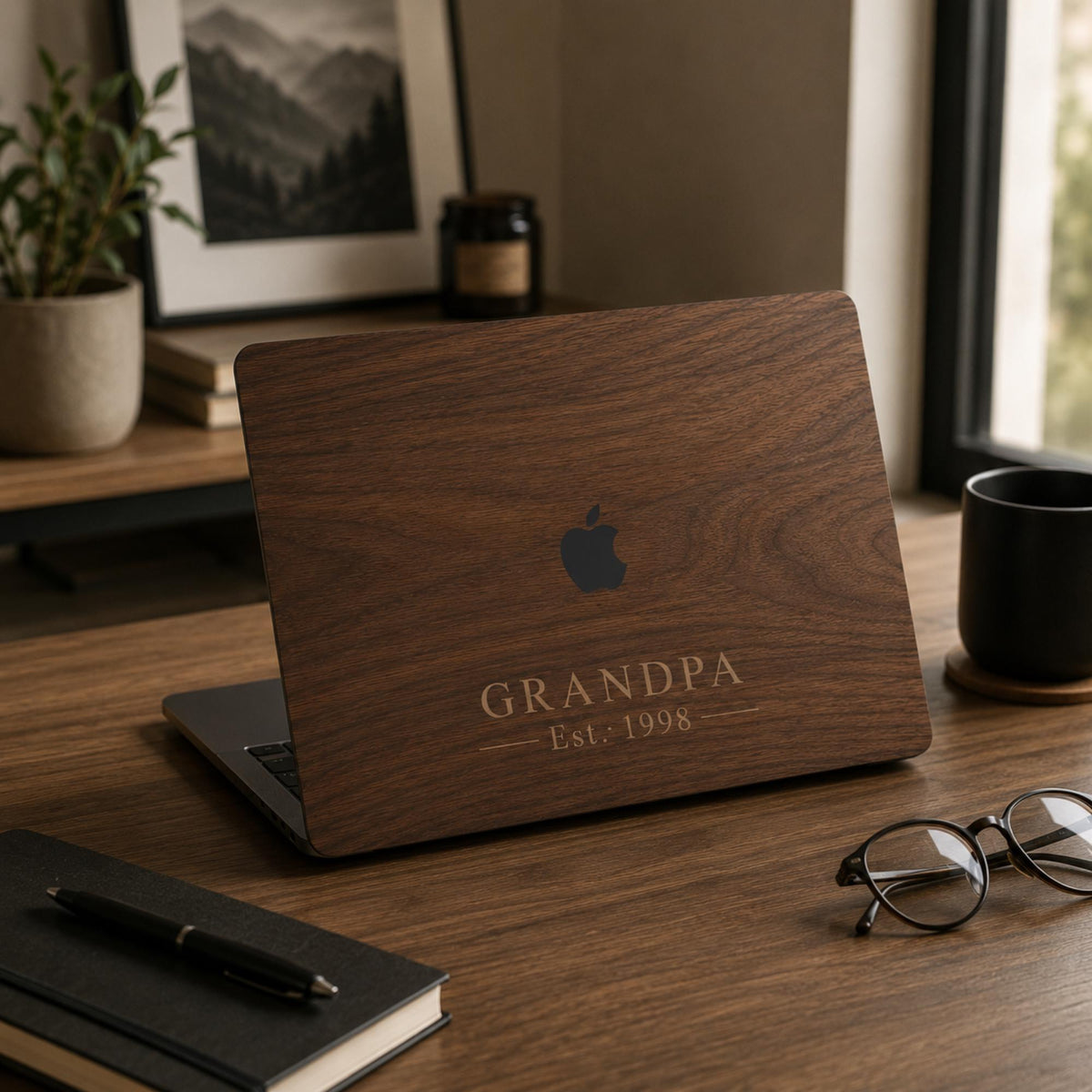 Personalized wood-look MacBook case with a warm walnut wood-grain print and the custom text GRANDPA Est. 1998 in elegant serif lettering, on a partially open laptop on a wooden desk with plants, books, glasses, and a coffee mug. The wood grain and lettering are a high-definition UV print on a smooth polycarbonate hard shell, not real wood. A heartfelt personalized gift for grandpa, Father's Day, or a milestone, fully customizable with your own word and year for any father figure.