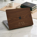 Personalized wood-look MacBook case with a warm walnut wood-grain print and the text DAD Est. 2026 in elegant serif lettering, displayed on a partially open laptop on a white marble desk with leather-bound books, a notebook, and a coffee cup. The wood grain and lettering are a high-definition UV print on a smooth polycarbonate hard shell, not real wood. A handsome personalized Father's Day, birthday, or new dad gift for him, customizable with your own word and year.