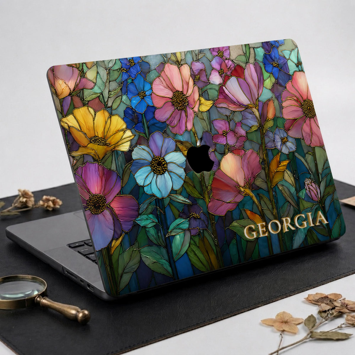 Personalized Colorful Garden MacBook case on dark leather with a brass magnifying glass and dried botanical stems.