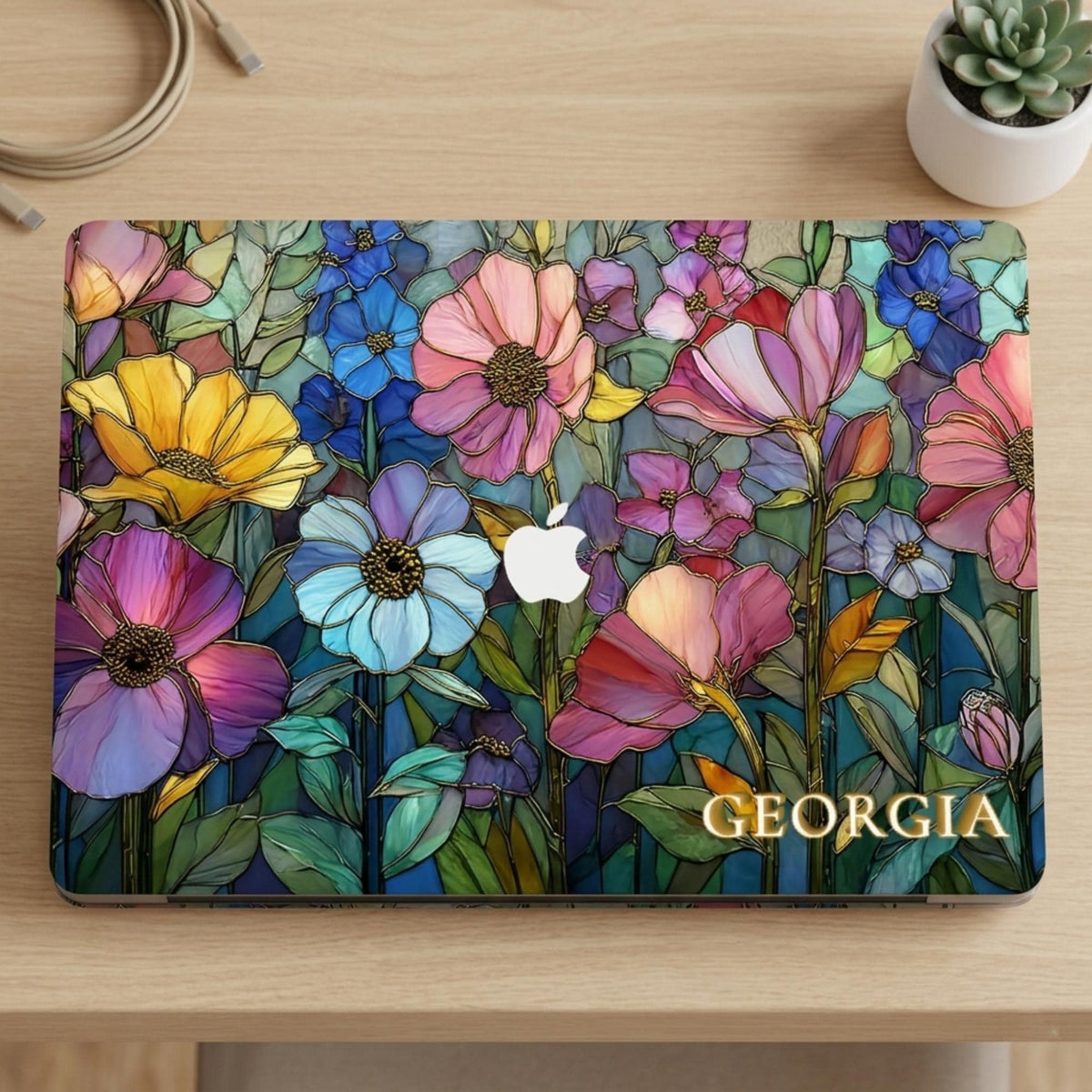 Flat lay of the Colorful Garden MacBook case with Georgia on light wood beside a white succulent pot and charging cable.