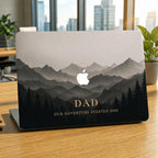 Personalized misty mountains MacBook case on a partially open laptop on a wooden desk by a window with a city view and plants. Layered mountain peaks fading into fog above a silhouette pine forest in soft grey and charcoal tones, with the custom text DAD Our Adventure Started 2020 and the Apple logo glowing among the peaks. The design is a high-definition UV print on a smooth polycarbonate hard shell. A personalized Father's Day adventure gift for him, customizable with your own words.