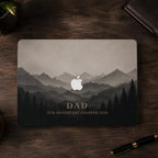 Top-down view of a personalized misty mountains MacBook case on a closed laptop on a dark wooden desk with a leather notebook and a fountain pen. Layered mountain peaks fading into fog above a silhouette pine forest in soft grey and charcoal tones, with the custom text DAD Our Adventure Started 2020 and the Apple logo glowing among the peaks. The design is a high-definition UV print on a smooth polycarbonate hard shell. A personalized Father's Day adventure gift, customizable with your words.