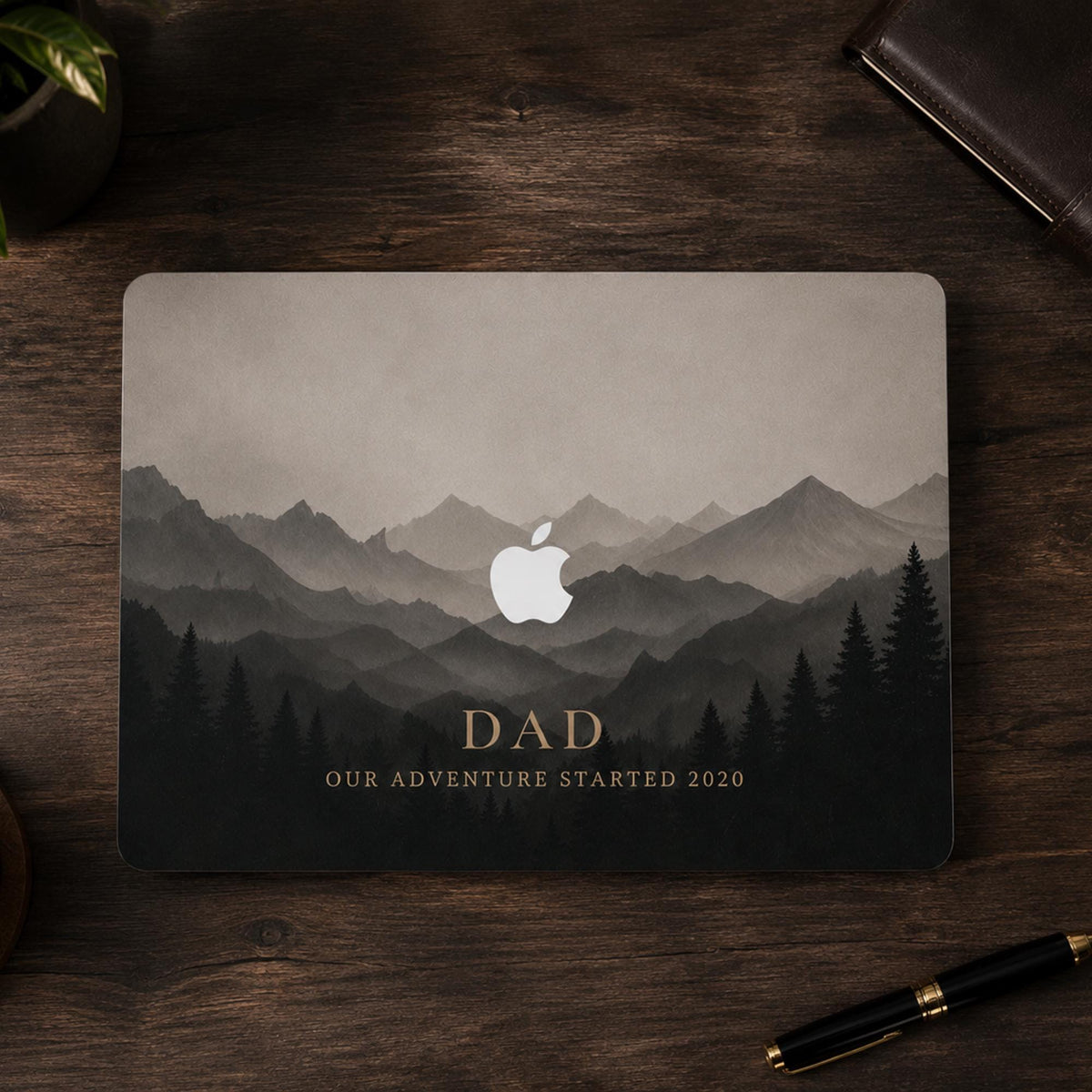 Top-down view of a personalized misty mountains MacBook case on a closed laptop on a dark wooden desk with a leather notebook and a fountain pen. Layered mountain peaks fading into fog above a silhouette pine forest in soft grey and charcoal tones, with the custom text DAD Our Adventure Started 2020 and the Apple logo glowing among the peaks. The design is a high-definition UV print on a smooth polycarbonate hard shell. A personalized Father's Day adventure gift, customizable with your words.