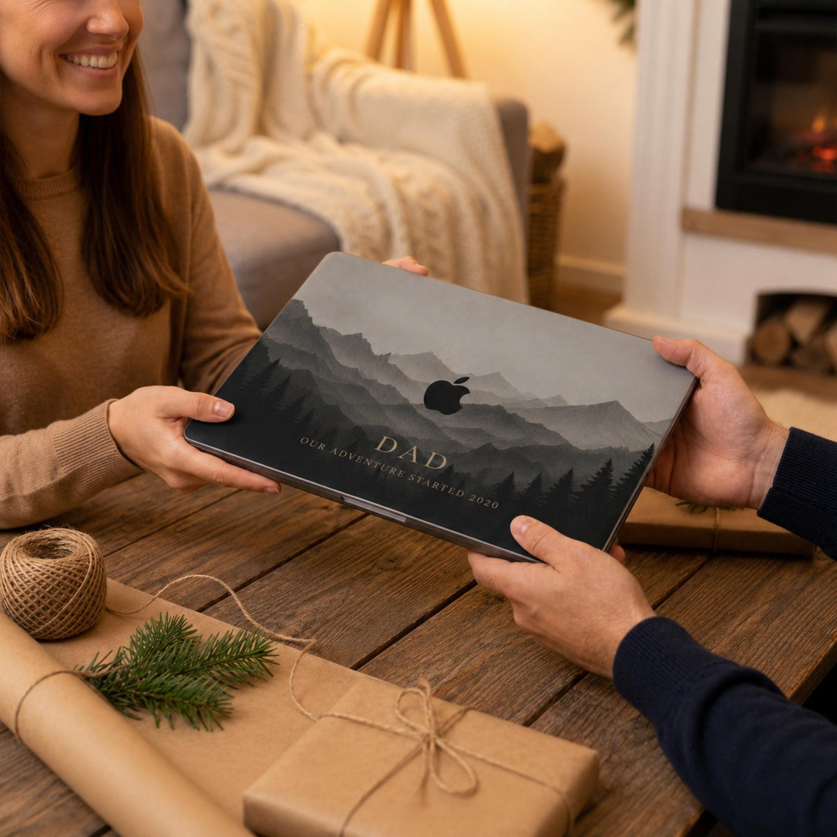 A woman handing a personalized misty mountains MacBook case as a gift to a man beside a fireplace, with rustic kraft gift wrapping and pine on the table. The case shows layered mountain peaks in soft grey and charcoal tones with the custom text DAD and the Apple logo among the peaks. The design is a high-definition UV print on a smooth polycarbonate hard shell. A heartfelt personalized Father's Day or Christmas gift for dad, customizable with your own words.