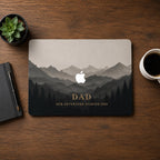 Personalized MacBook case with misty mountain landscape design featuring layered dark forest silhouettes and soft foggy peaks fading into warm beige sky. Custom text DAD and phrase Our Adventure Started 2020 displayed in elegant gold lettering at bottom. Dark charcoal and warm taupe tones with cinematic depth. Shown flat lay on dark wooden desk with black notebook, pen, succulent plant, and coffee cup. Perfect personalized gift for dad, husband, and outdoor adventure enthusiasts.