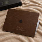 Personalized wood-look MacBook case with a rich walnut wood-grain print and the text DAD Est. 2026 in clear elegant serif lettering, shown on a closed laptop on a soft beige blanket. The wood grain and lettering are a high-definition UV print on a smooth polycarbonate hard shell, not real wood. A handsome personalized Father's Day, birthday, or new dad gift for him, customizable with your own word and year, beautifully detailed and quietly masculine on any desk.