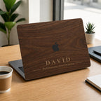 Personalized dark brown wood grain effect MacBook case from TrioAuraDesign, shown on a modern desk with custom name “DAVID” and a short personalized phrase below. The smooth hard shell features a realistic walnut wood look with the Apple logo visible at center. The wood grain and lettering are professionally printed, not real wood or engraving. Customizable for compatible MacBook Air, Pro and Neo models.