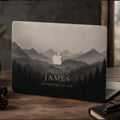 Personalized misty mountains MacBook case showing the custom text JAMES Adventure Awaits, with layered mountain peaks fading into fog above a silhouette pine forest in soft grey and charcoal tones, the Apple logo glowing among the peaks. Shown on a partially open laptop on a dark wooden desk with pine and a mug by a window. The design is a high-definition UV print on a smooth polycarbonate hard shell. A personalized adventure, birthday, or graduation gift, customizable with any name.