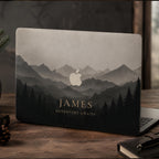 Personalized misty mountains MacBook case showing the custom text JAMES Adventure Awaits, with layered mountain peaks fading into fog above a silhouette pine forest in soft grey and charcoal tones, the Apple logo glowing among the peaks. Shown on a partially open laptop on a dark wooden desk with pine and a mug by a window. The design is a high-definition UV print on a smooth polycarbonate hard shell. A personalized adventure, birthday, or graduation gift, customizable with any name.