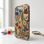 Botanical embroidery effect iPhone case standing upright on a white marble surface beside delicate wildflowers and a vintage magnifying glass. The cream-colored design features detailed printed embroidery-style wildflowers, ferns, a butterfly, bumblebee, and hummingbird, creating the look of handcrafted stitching. The case has a smooth glossy finish with a black protective bumper and precise cutouts, photographed in soft natural daylight. Available from TrioAuraDesign.