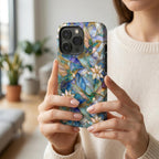 Woman holding a stained glass effect iPhone case with colorful hummingbirds, white lilies, and botanical artwork inspired by classic stained glass windows. The glossy protective case features vibrant blue, teal, ivory, and gold tones with smooth edge-to-edge UV printing. Photographed in soft natural daylight to showcase the elegant artistic design and everyday use. Available from TrioAuraDesign.