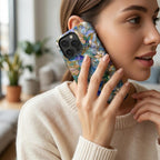 Woman holding a stained glass effect iPhone case to her ear during a phone call in a bright modern home. The glossy protective case features colorful hummingbirds, white lilies, and botanical artwork in rich blue, teal, ivory, and gold tones inspired by classic stained glass windows. The printed design wraps beautifully around the case, creating an elegant artistic look while showcasing everyday use. Available from TrioAuraDesign.