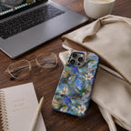 Stained glass effect MagSafe-compatible iPhone case featuring vibrant hummingbirds, elegant white lilies, and botanical artwork in rich blue, teal, ivory, and gold tones displayed on a stylish wooden desk beside a laptop, notebook, eyeglasses, coffee, and canvas tote bag. The glossy protective case showcases a smooth printed stained glass effect with premium edge-to-edge UV printing, creating an elegant everyday workspace aesthetic. Available from TrioAuraDesign.
