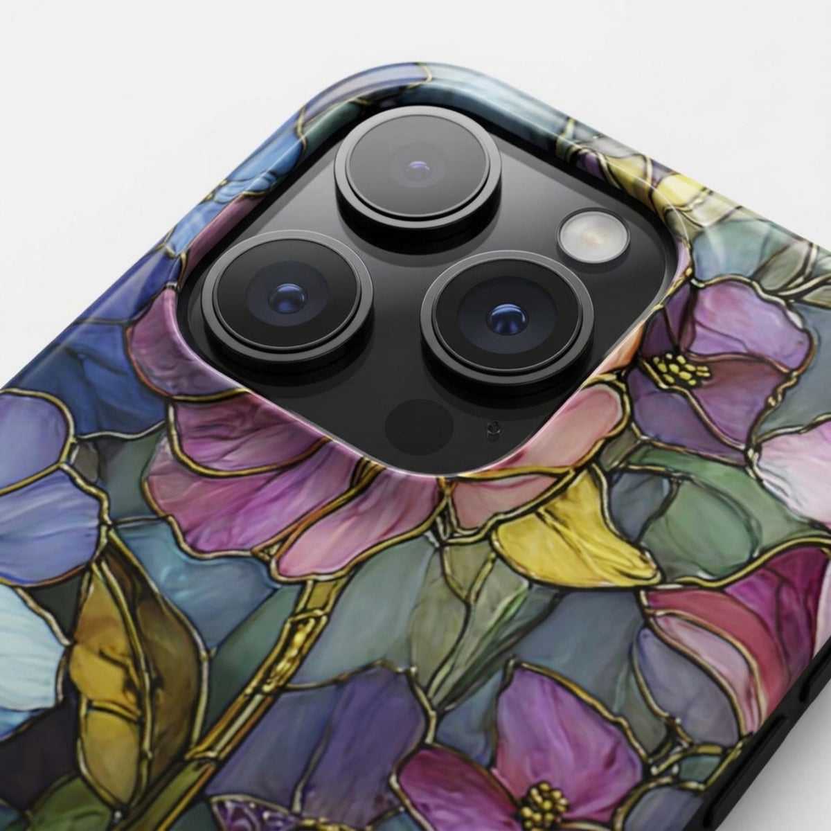 Close-up detail of a colorful stained glass effect iPhone case featuring layered purple, pink, blue, green and yellow botanical artwork with intricate golden-looking printed outlines. The macro product view highlights the raised protective camera bezel, precise camera opening, full-wrap print and smooth glossy surface. The stained glass appearance is a printed visual effect with no real glass, metal or raised decorative texture, available from TrioAuraDesign.