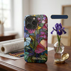 Personalized iPhone case with the custom name Georgia, featuring a colorful stained glass effect with layered pink, purple, blue and green flowers, leafy details and intricate golden-looking printed outlines. Displayed upright on a wooden desk beside an iris flower, art supplies and a compass, the product-focused scene highlights the personalization, botanical artwork and protective camera area. The stained glass appearance is a printed visual effect on a smooth glossy case from TrioAuraDesign.