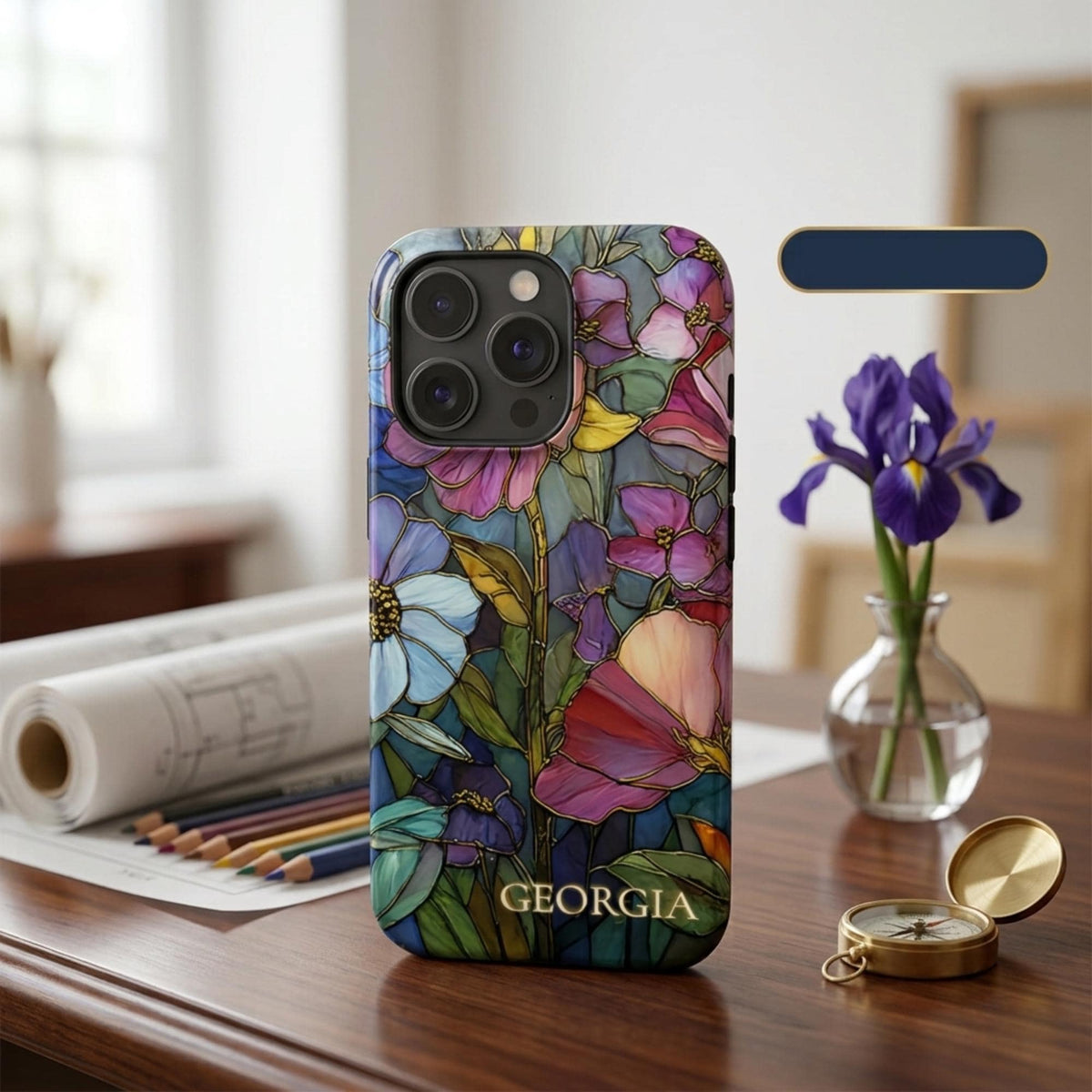 Personalized iPhone case with the custom name Georgia, featuring a colorful stained glass effect with layered pink, purple, blue and green flowers, leafy details and intricate golden-looking printed outlines. Displayed upright on a wooden desk beside an iris flower, art supplies and a compass, the product-focused scene highlights the personalization, botanical artwork and protective camera area. The stained glass appearance is a printed visual effect on a smooth glossy case from TrioAuraDesign.