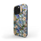 Angled view of a stained glass effect iPhone case featuring colorful hummingbirds, white lilies, and botanical artwork in rich blue, teal, ivory, and gold tones. The glossy protective case showcases smooth edge-to-edge UV printing inspired by classic stained glass windows while revealing the slim profile, rounded edges, and side button design. Displayed on a clean white background. Available from TrioAuraDesign.